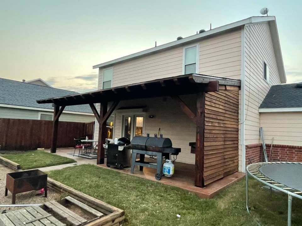 Backyard patio with a covered grill area beside a two-story house and fenced lawn.