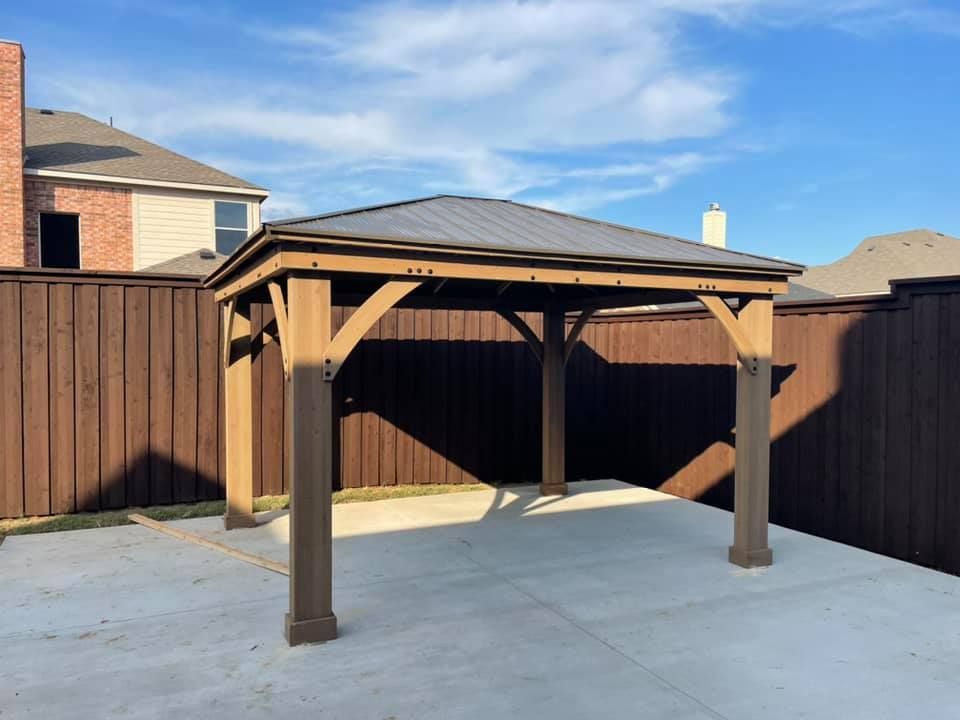 Wooden backyard pergola with a dark roof on a concrete patio, bordered by tall brown fences