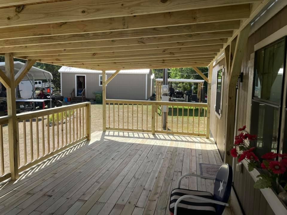 Covered wooden porch with railing, ceiling beams, and a chair beside a house window, facing a grassy yard.