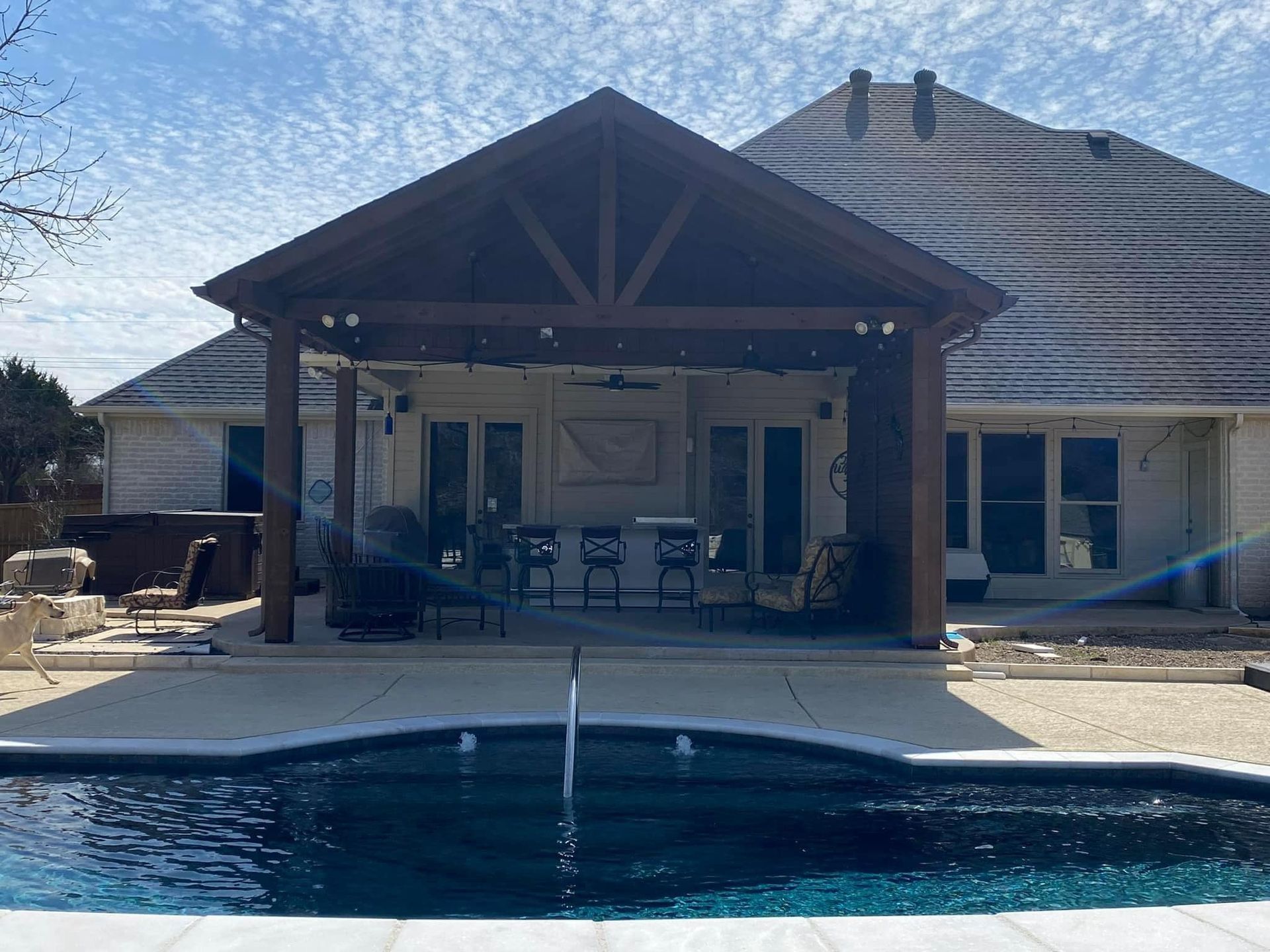 Backyard pool with a covered patio and fountain, facing a house on a sunny day