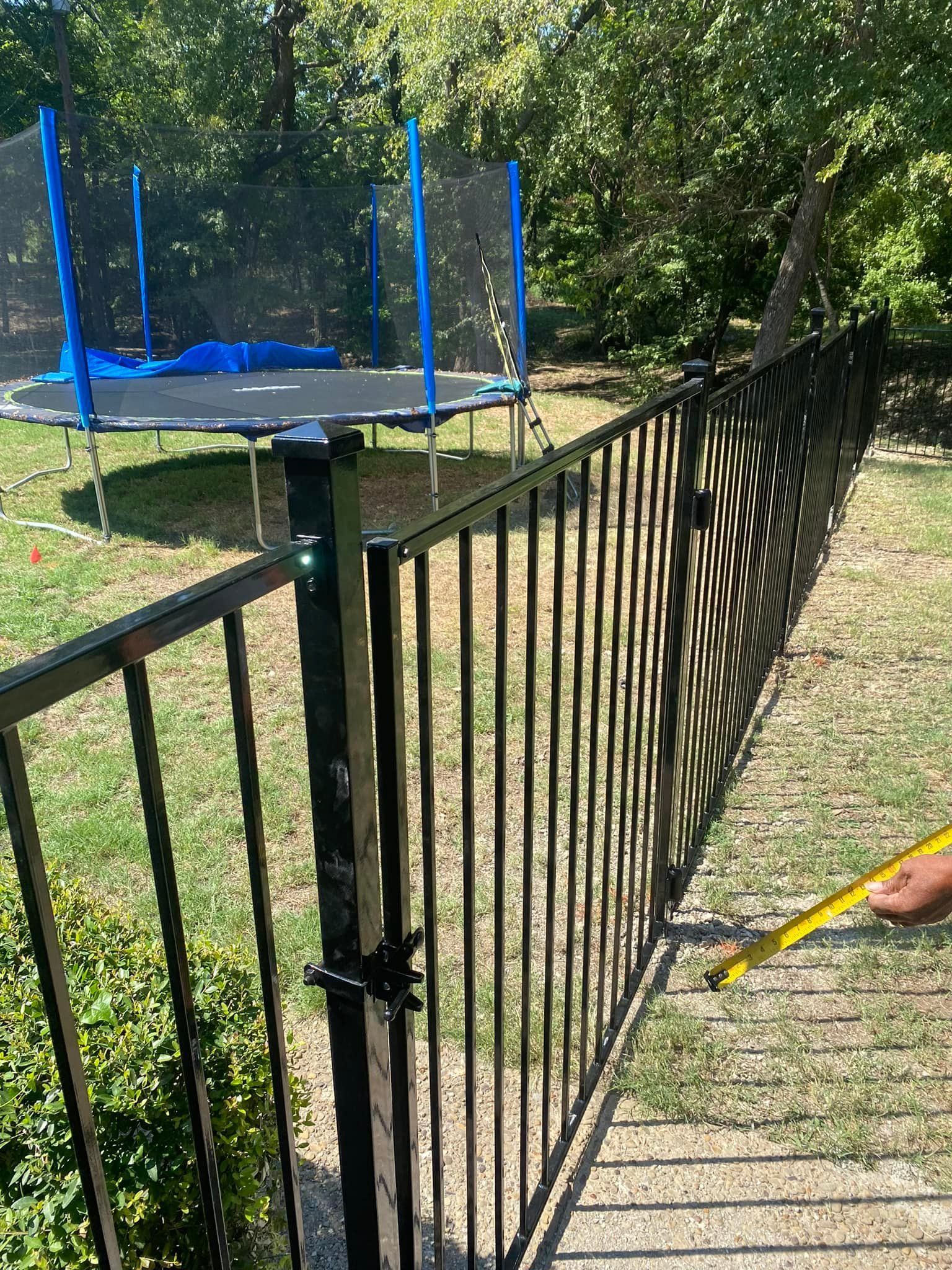 Black metal fence beside a blue trampoline in a grassy backyard with trees and a paved path