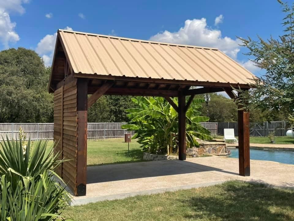 Wooden park pavilion with tan metal roof, set on a concrete pad in a grassy outdoor area.