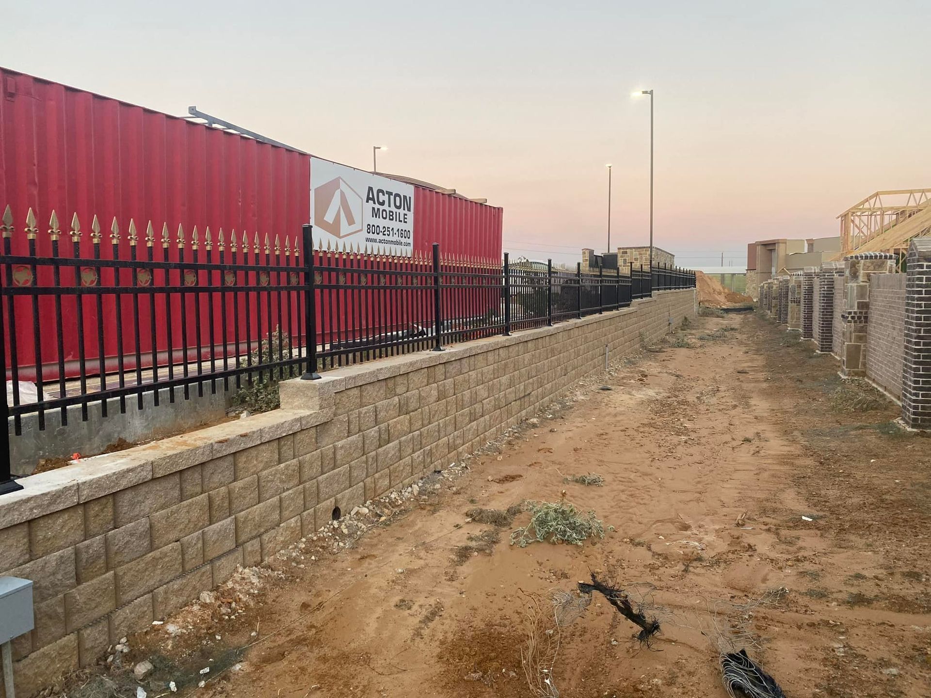 Dirt path beside a black fence and red shipping containers at a rail yard at sunset