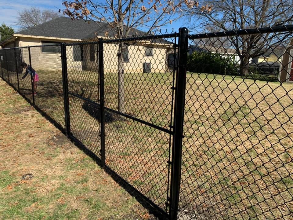 Black chain-link fence beside a tan house in a grassy yard on a sunny day