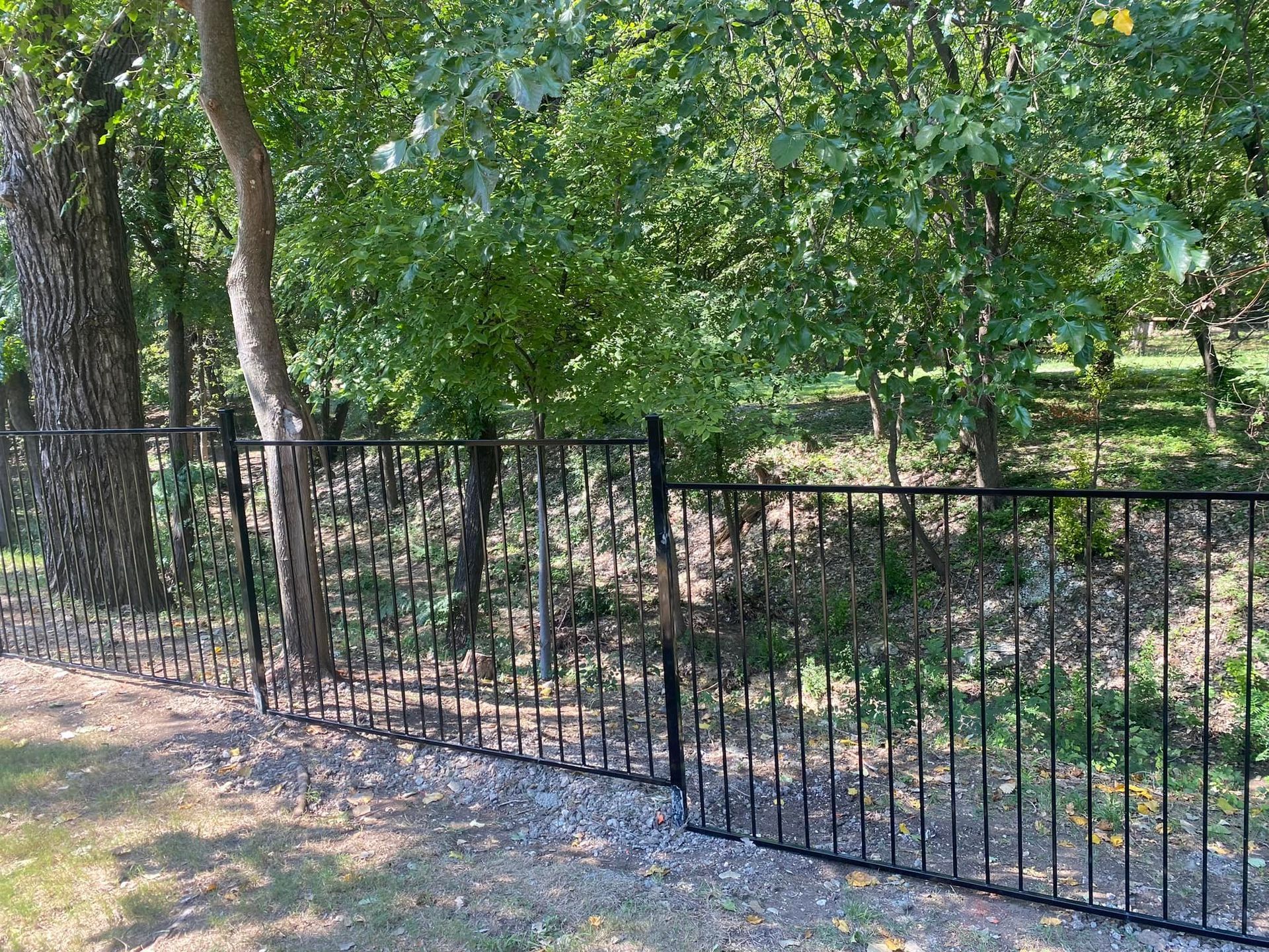 Black metal fence along a gravel path beside a wooded area and green trees