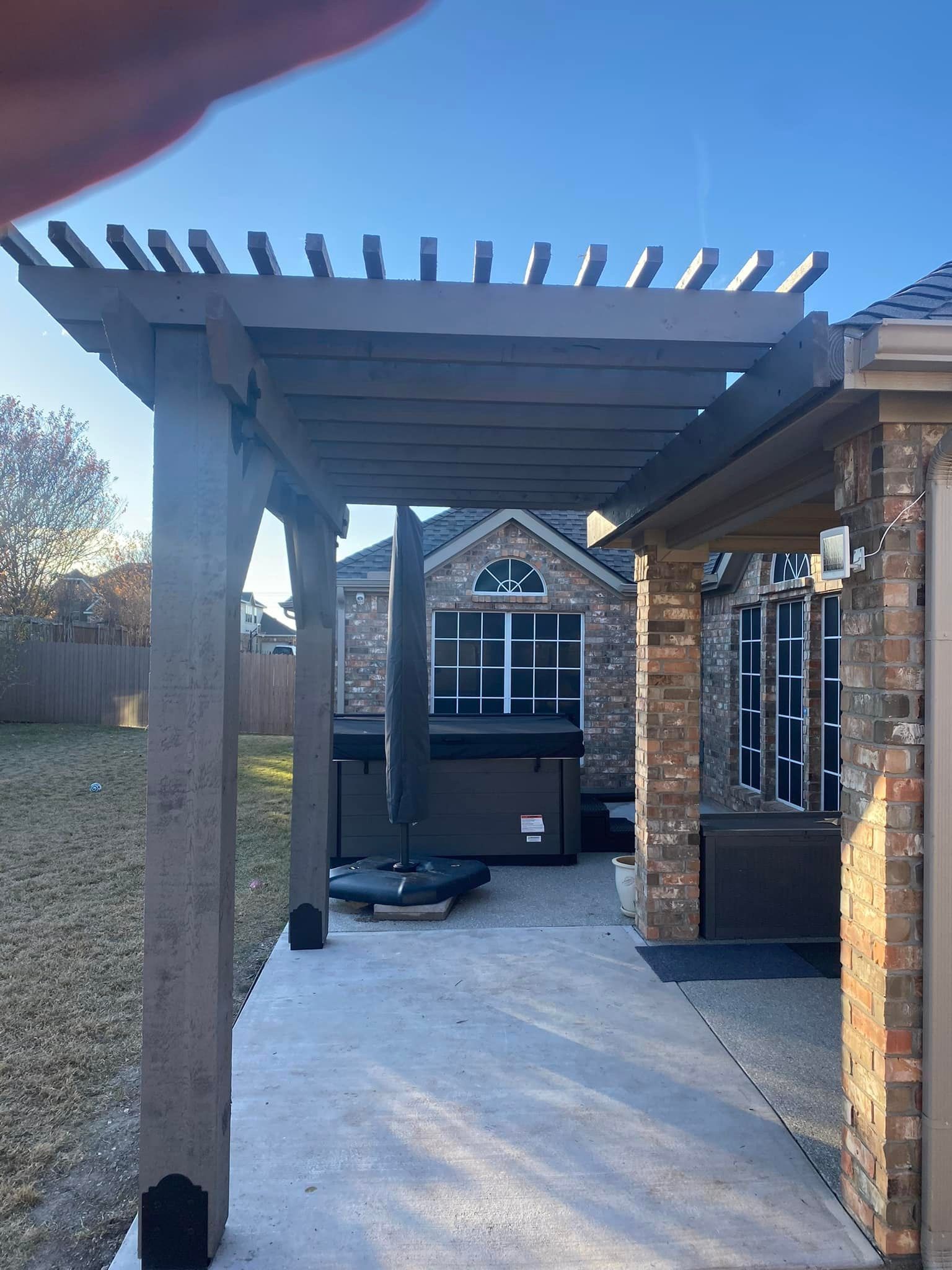 Covered outdoor patio with a pergola, brick house, and concrete walkway leading to a grill area.