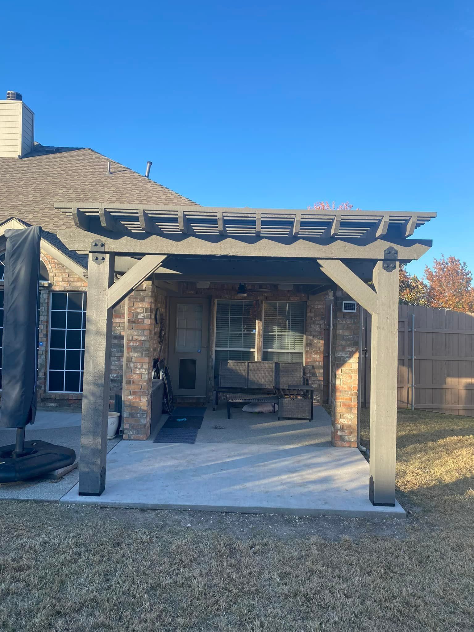 Stone patio with a wooden pergola and outdoor seating beside a house, under a clear blue sky