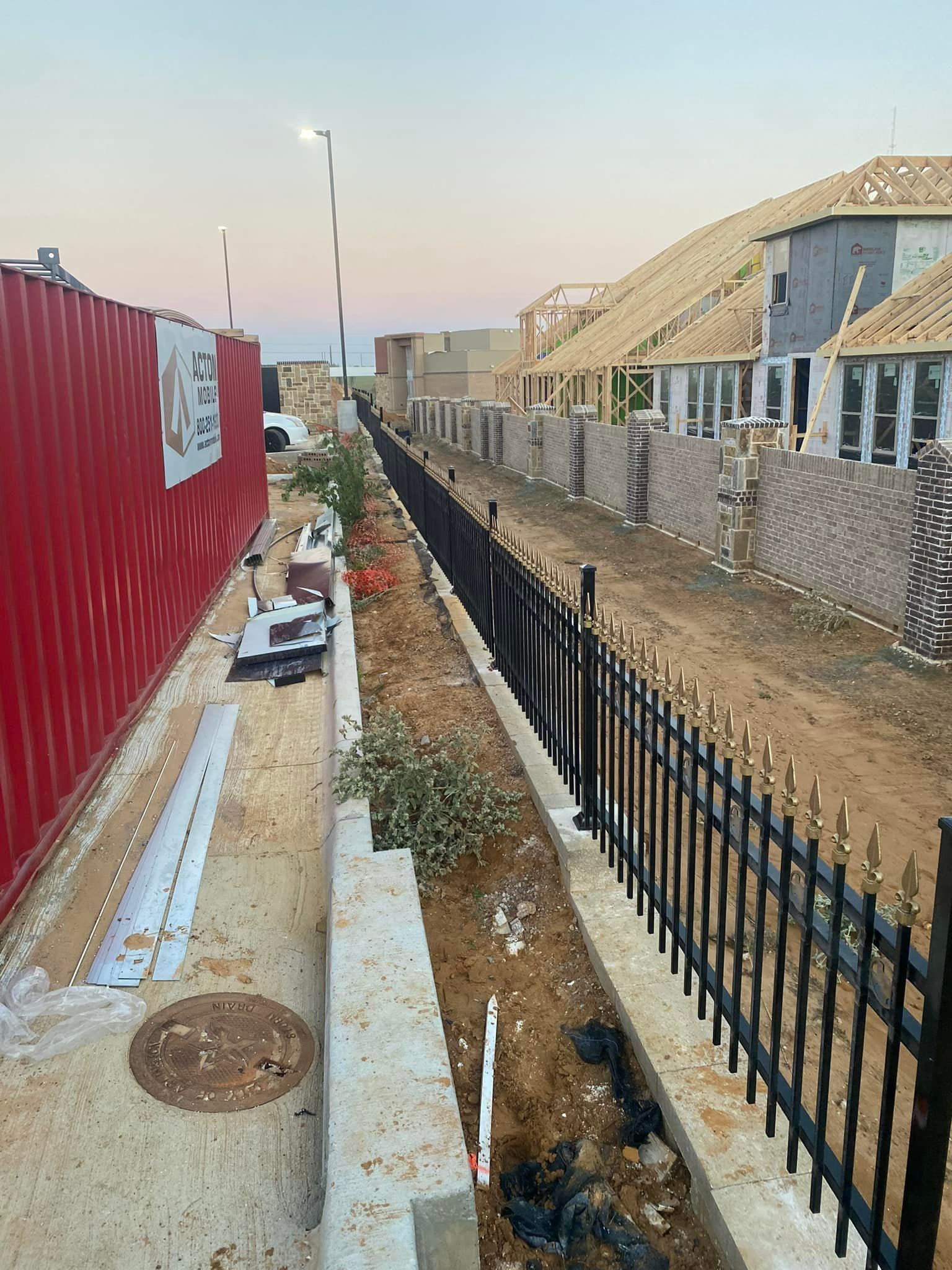 Construction site beside a black fence, with red barriers on the left and unfinished buildings on the right.