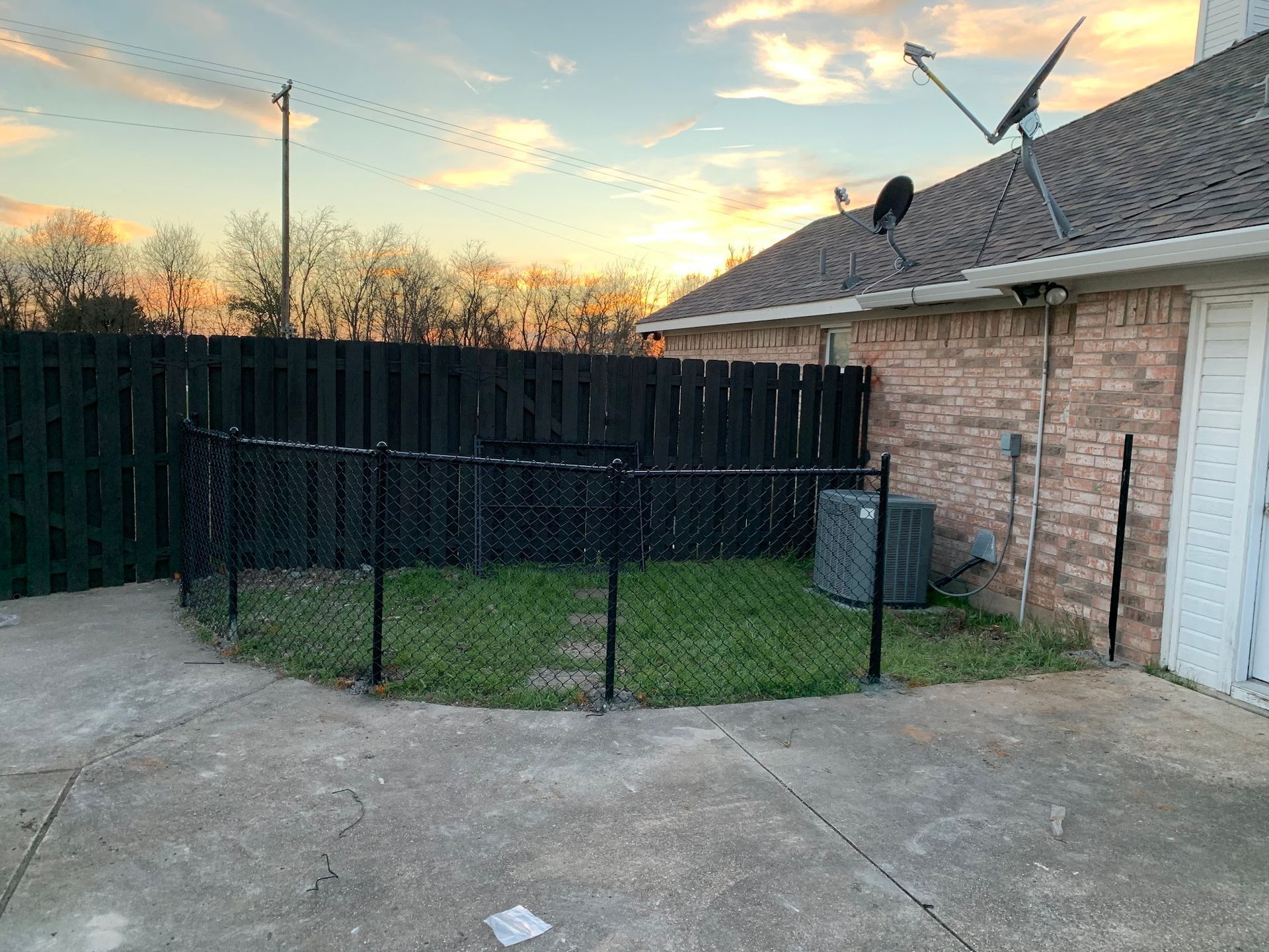 Backyard with black fenced grass enclosure, concrete patio, and house roof at sunset