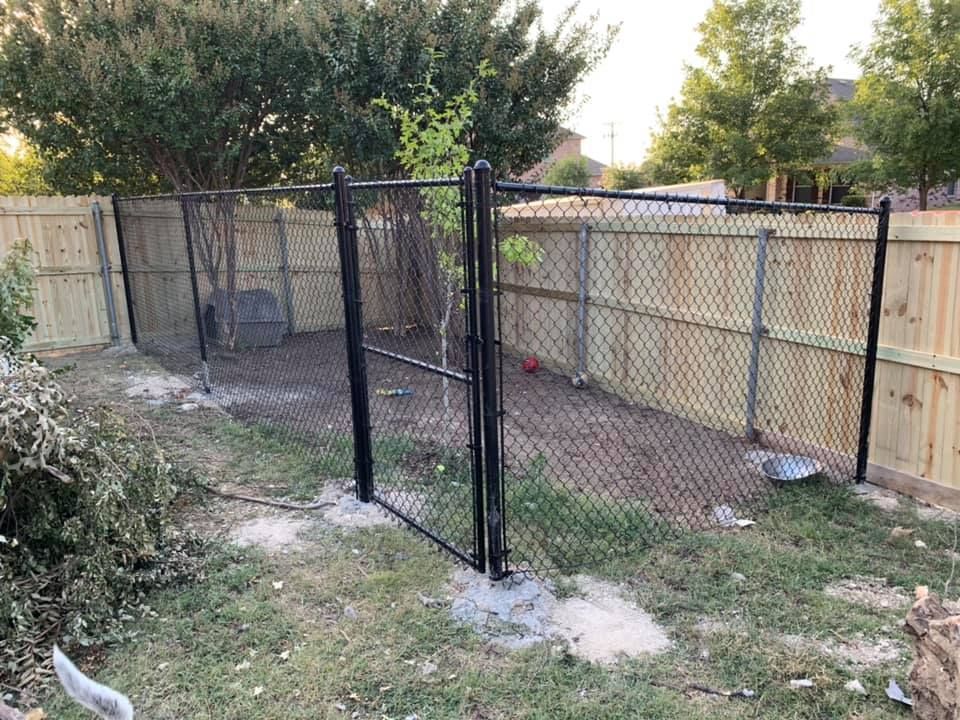 Small fenced backyard with a black chain-link gate and wooden privacy fence, bordered by grass and trees.