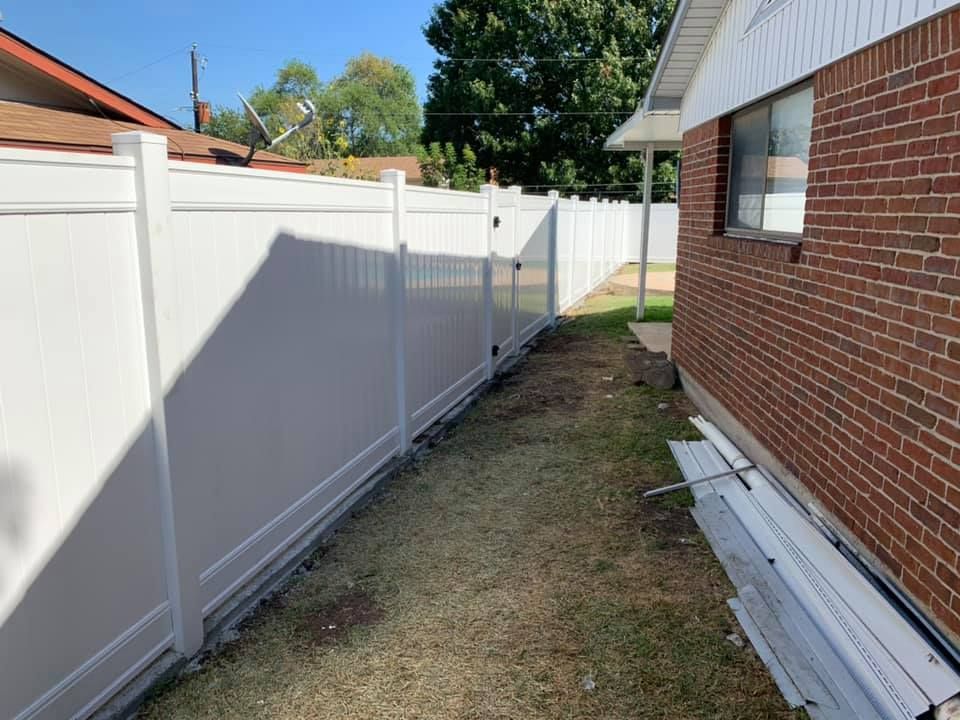 Narrow side yard between a white fence and brick house, with stacked siding panels along the wall.