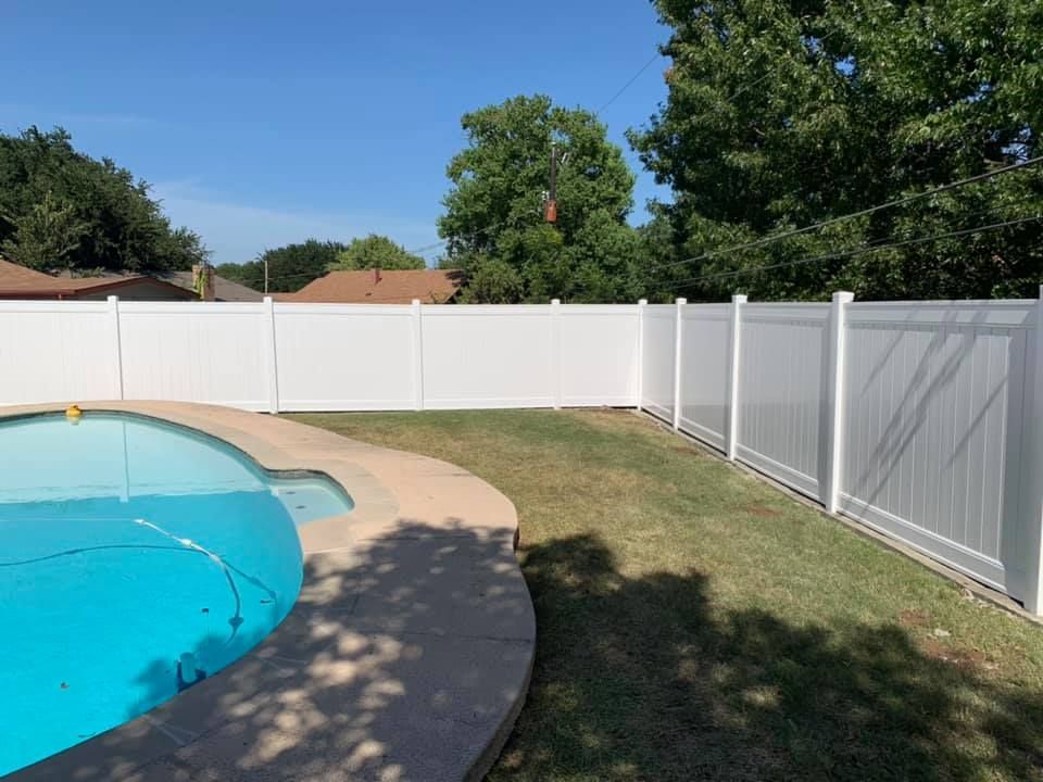 Backyard pool with curved deck, white privacy fence, green lawn, and trees under a blue sky