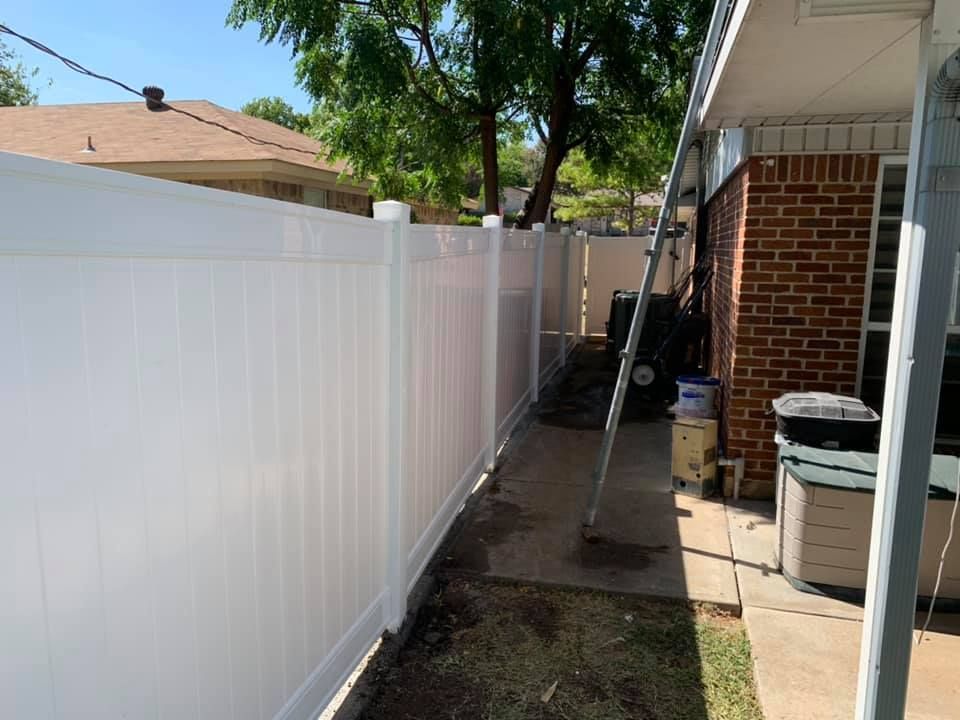 Narrow backyard side yard with white fence, brick house, and a garden hose beside bins.