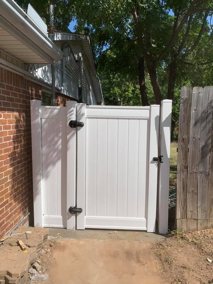 White wooden gate between a brick house and fence, opening to a dirt path in a shaded yard.