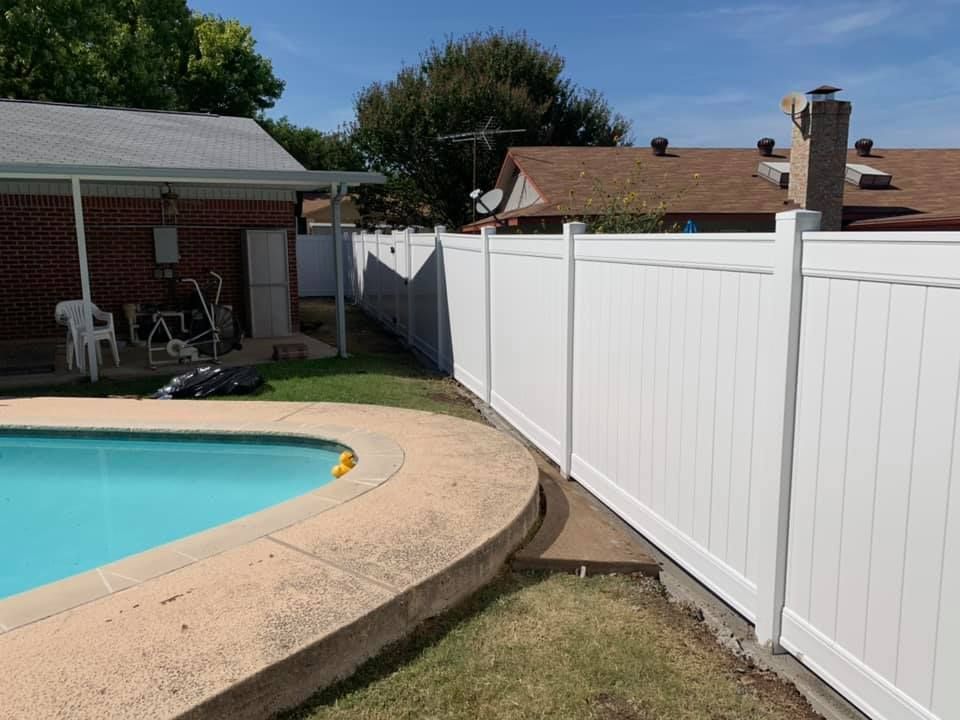 Backyard pool with curved stone deck and white privacy fence beside a brick house