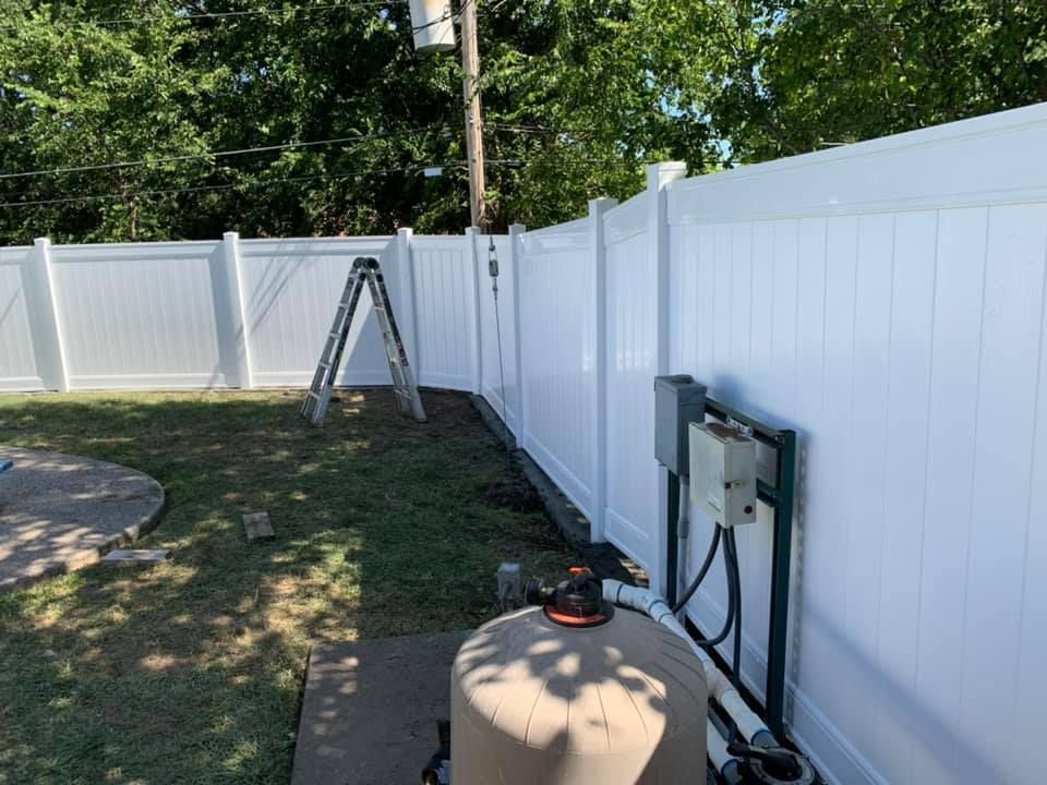 White vinyl fence and backyard pool equipment beside a beige tank, with a ladder in the grassy yard.