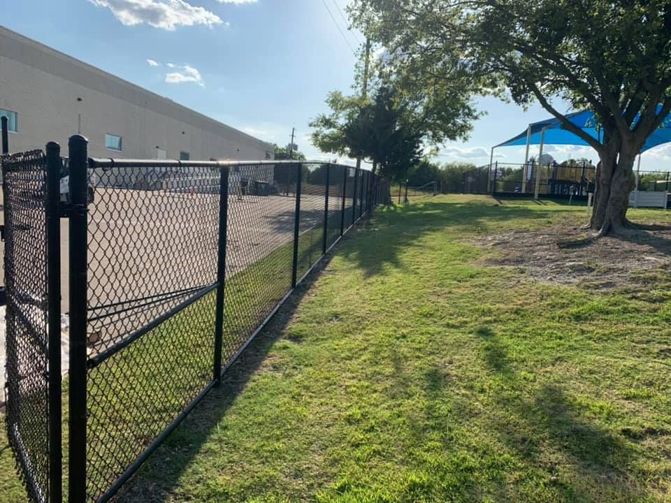 Chain-link fence beside a building, with grass and trees along a sunny path.