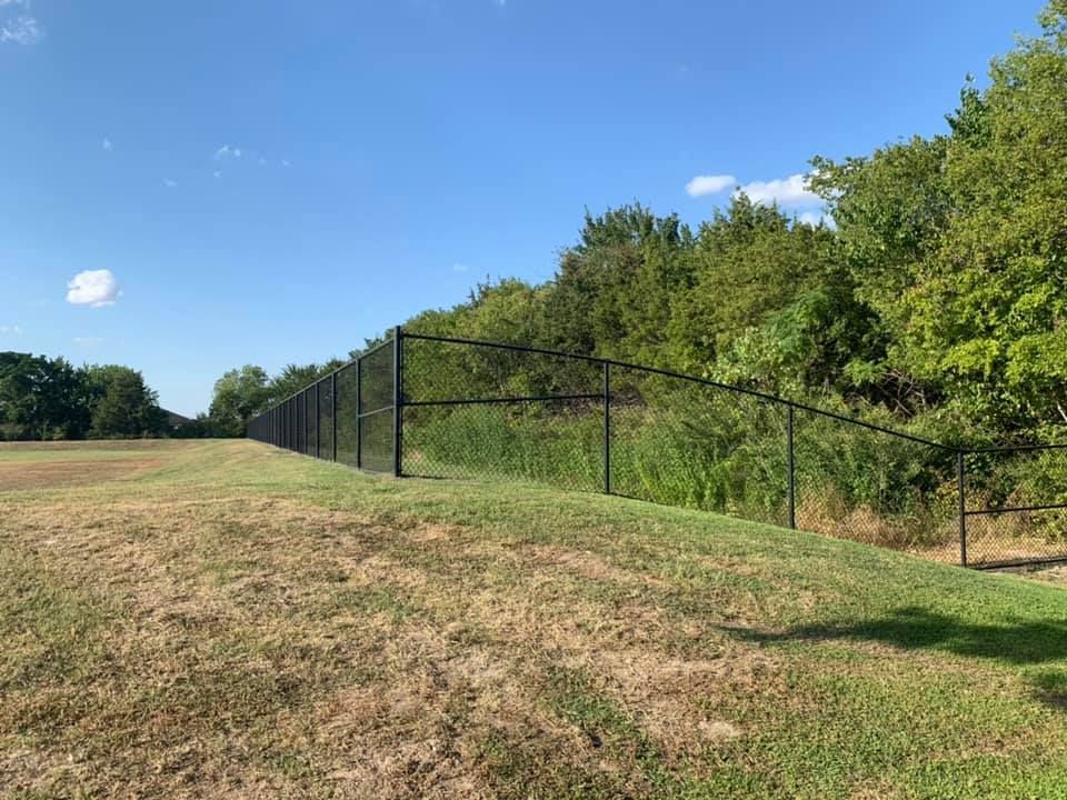 Long black chain-link fence along a grassy field beside green trees under a blue sky
