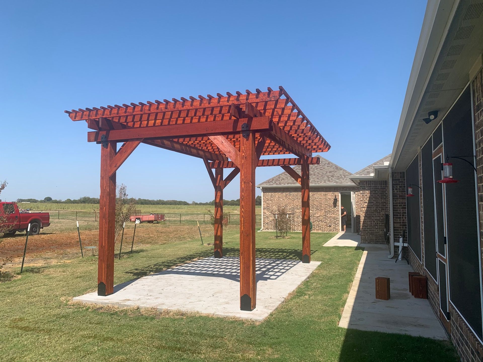 Red wooden pergola over gravel beside a house in a sunny rural yard