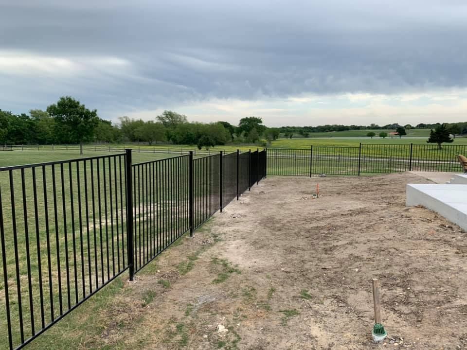 Black metal fence along a dirt yard beside a grassy field under cloudy skies.