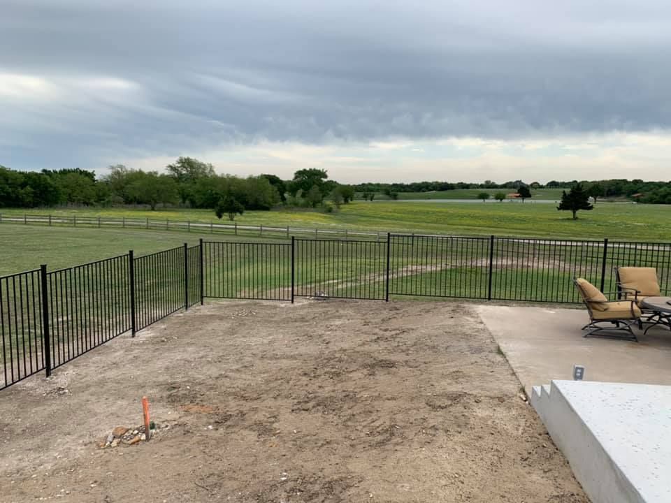 Backyard patio with black fence, tan lounge chairs, and a grassy field beyond under a cloudy sky