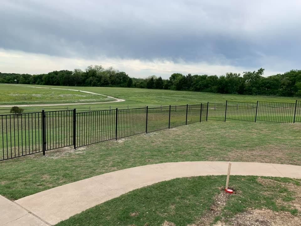 Grassy sports field with black fence, curving sidewalk, and cloudy sky.