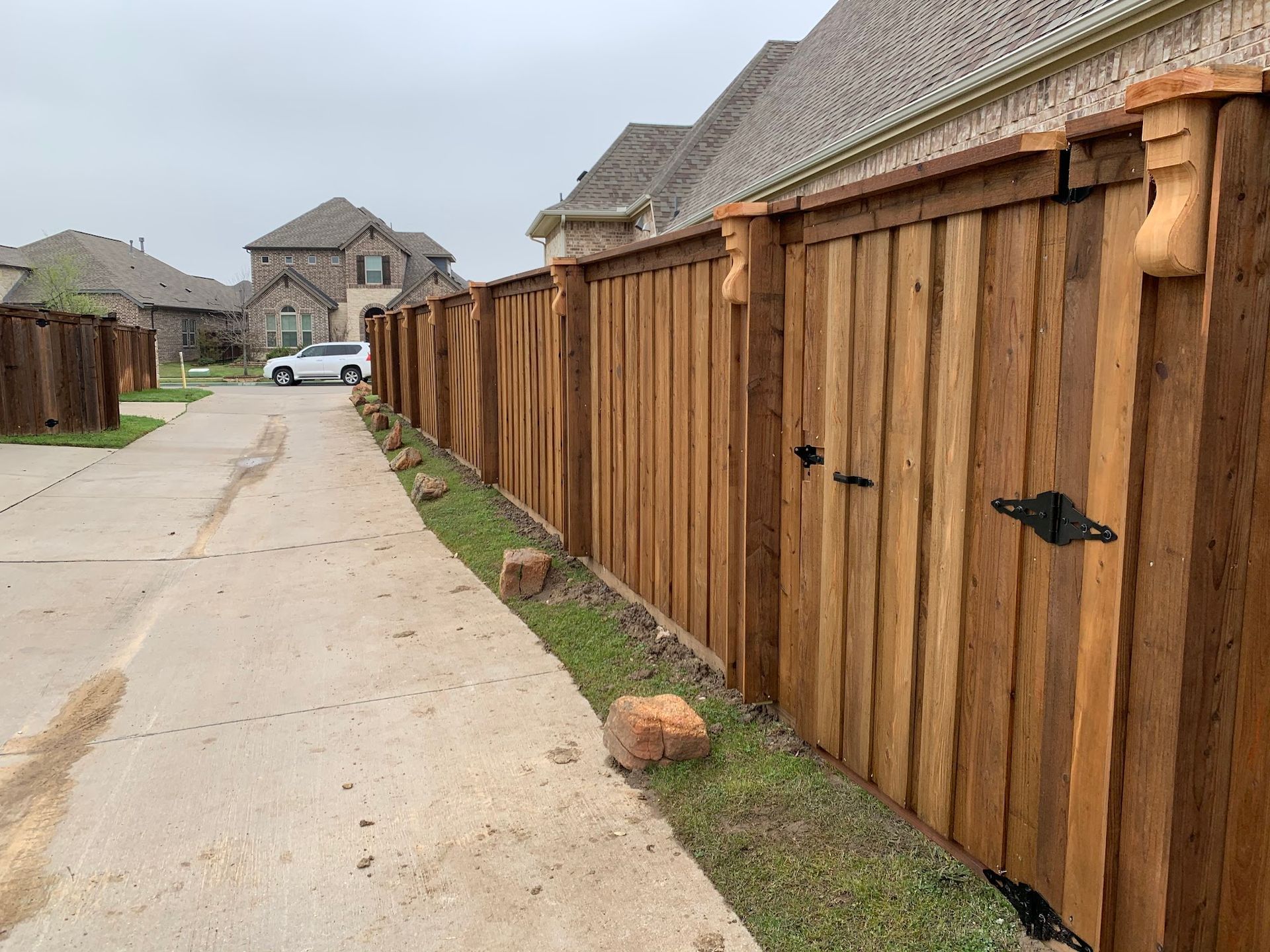 Wooden privacy fence along a concrete driveway in a suburban neighborhood, with houses in the background.