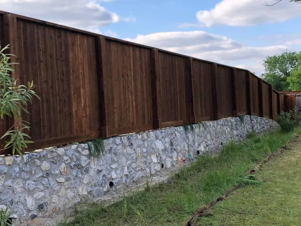 Wooden privacy fence atop a stone retaining wall beside a grassy path under a cloudy sky