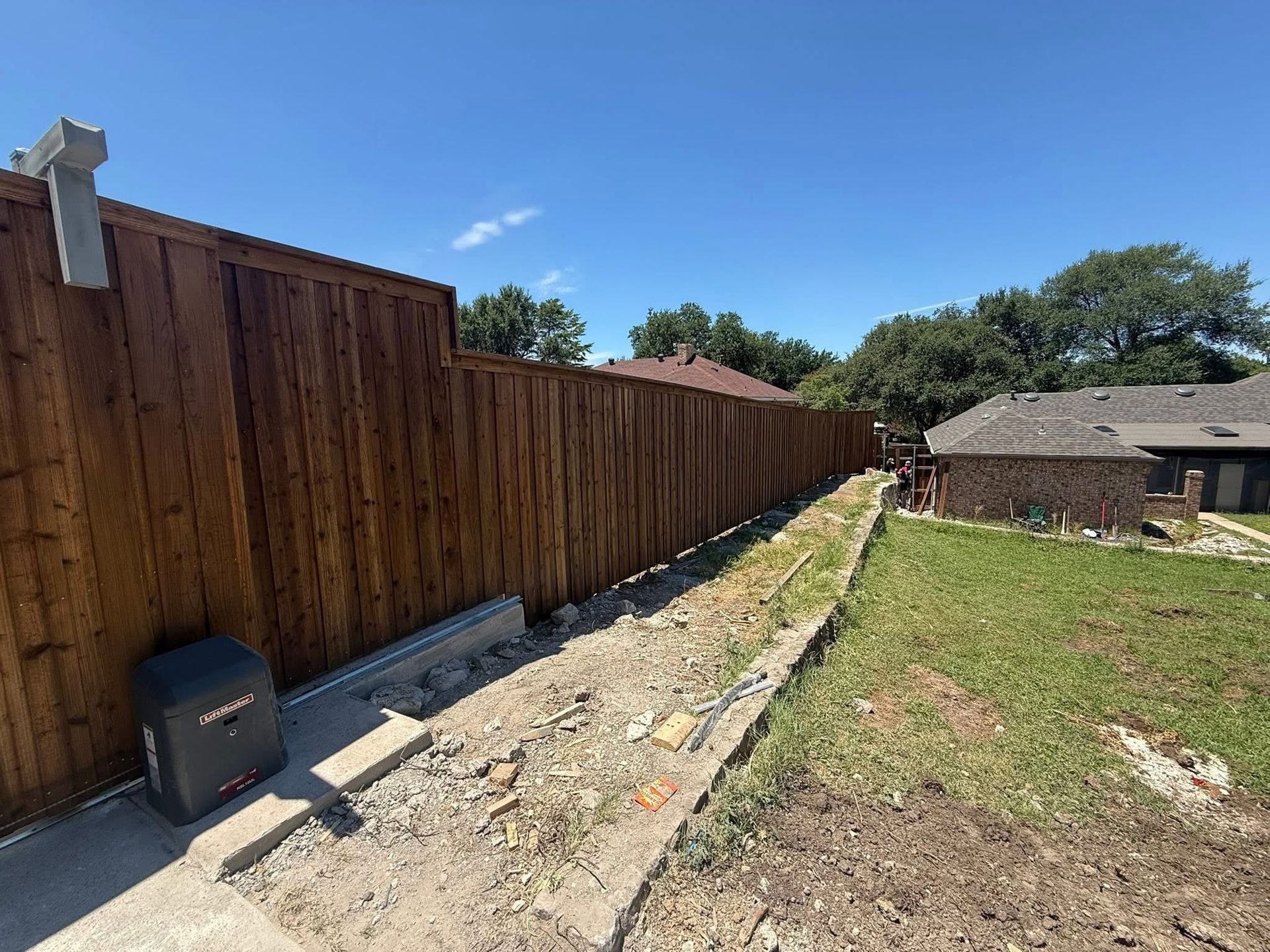 Wooden privacy fence along a narrow dirt strip beside a grassy yard and a house under a blue sky