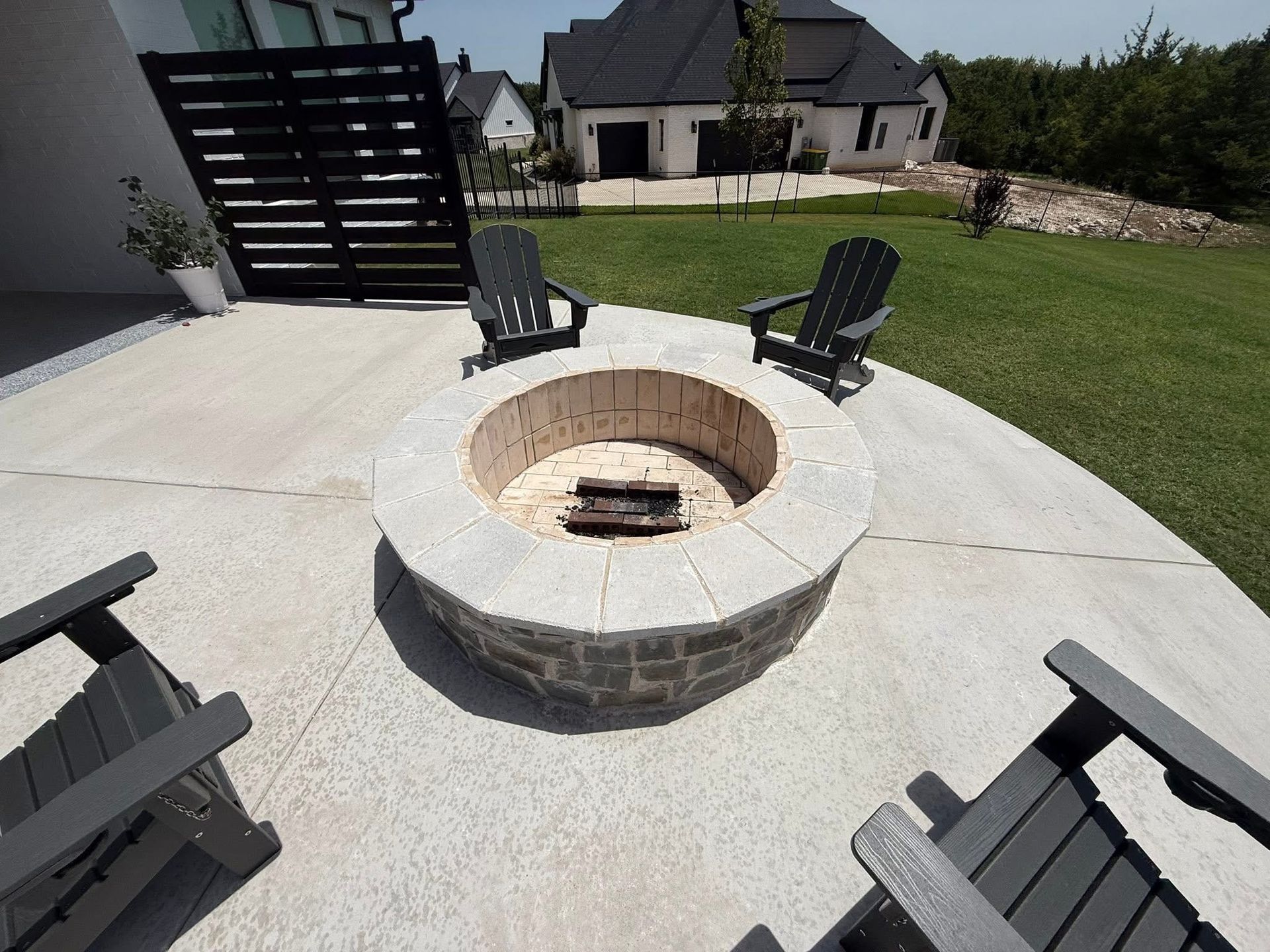 Backyard patio with a circular stone fire pit and four black chairs on a concrete surface