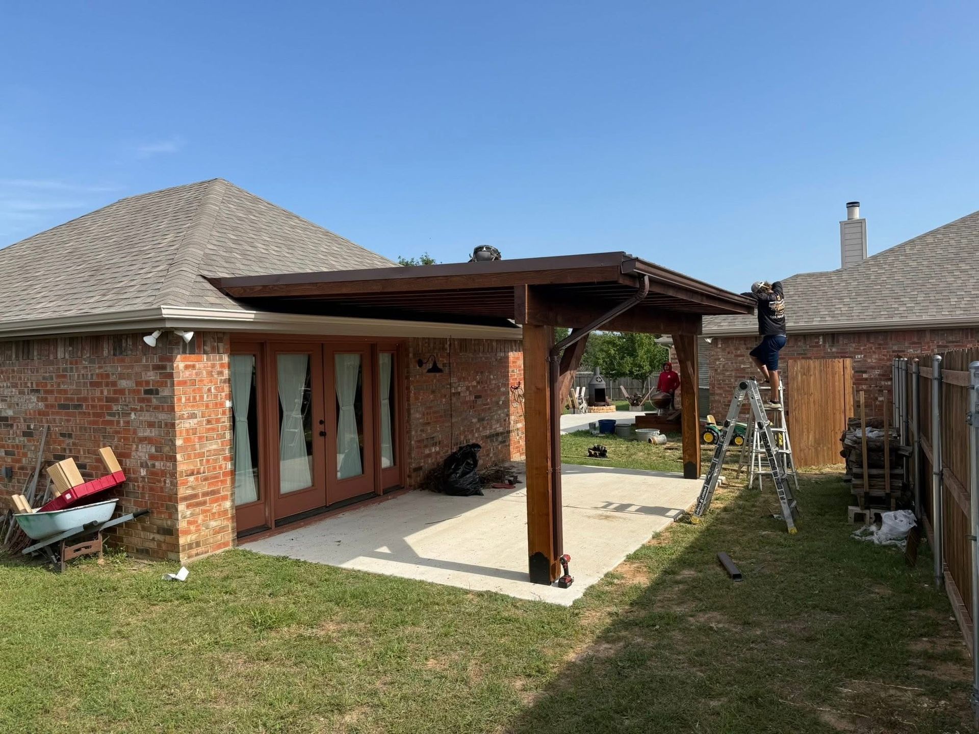 Backyard patio with a wooden pergola, brick house, concrete slab, and ladder near the fence.