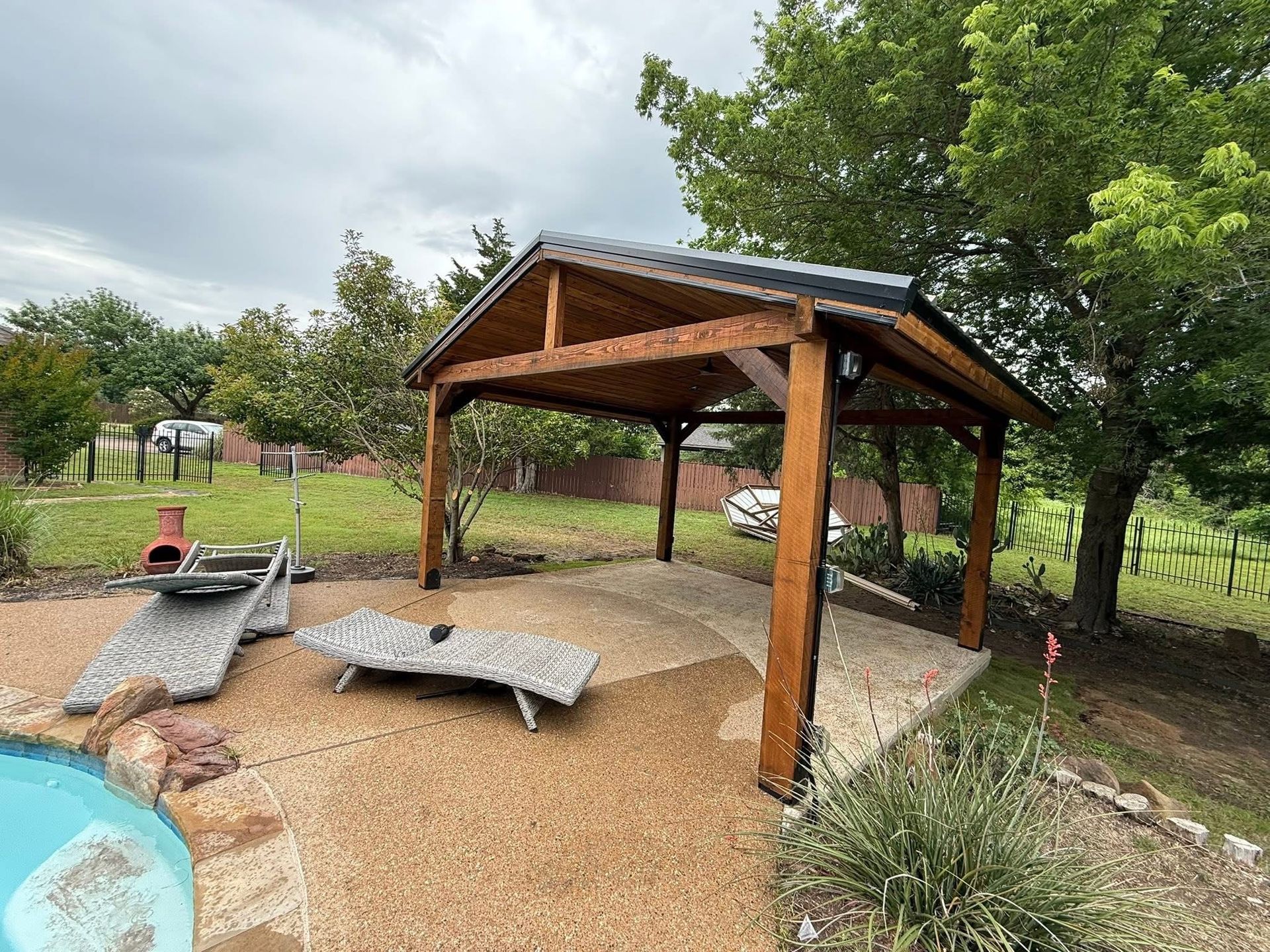 Wooden pavilion with benches on gravel in a grassy park, surrounded by trees and landscaping