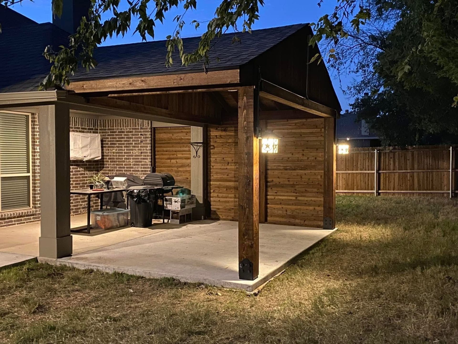 Backyard patio with a wooden pergola, lit lamp, grill, and fenced yard at dusk