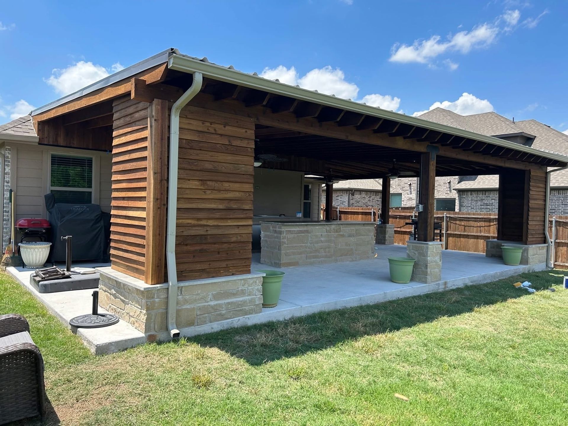 Wooden pavilion with a covered patio, stone base, and grassy yard under a blue sky
