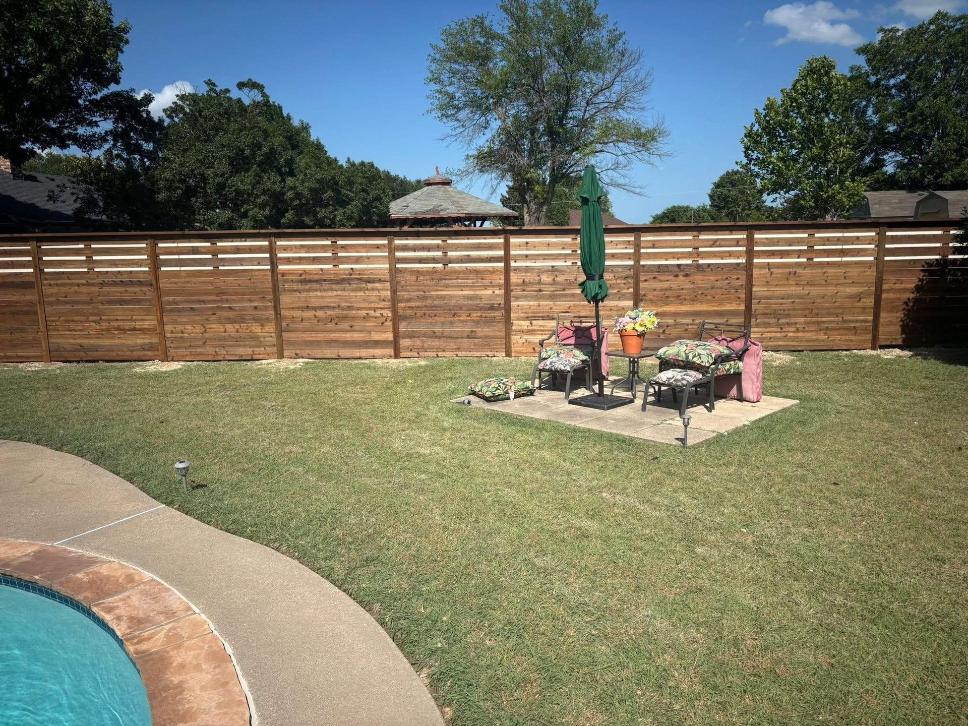 Backyard patio with pool, lawn, wooden fence, and a small table with colorful flowers under a tree