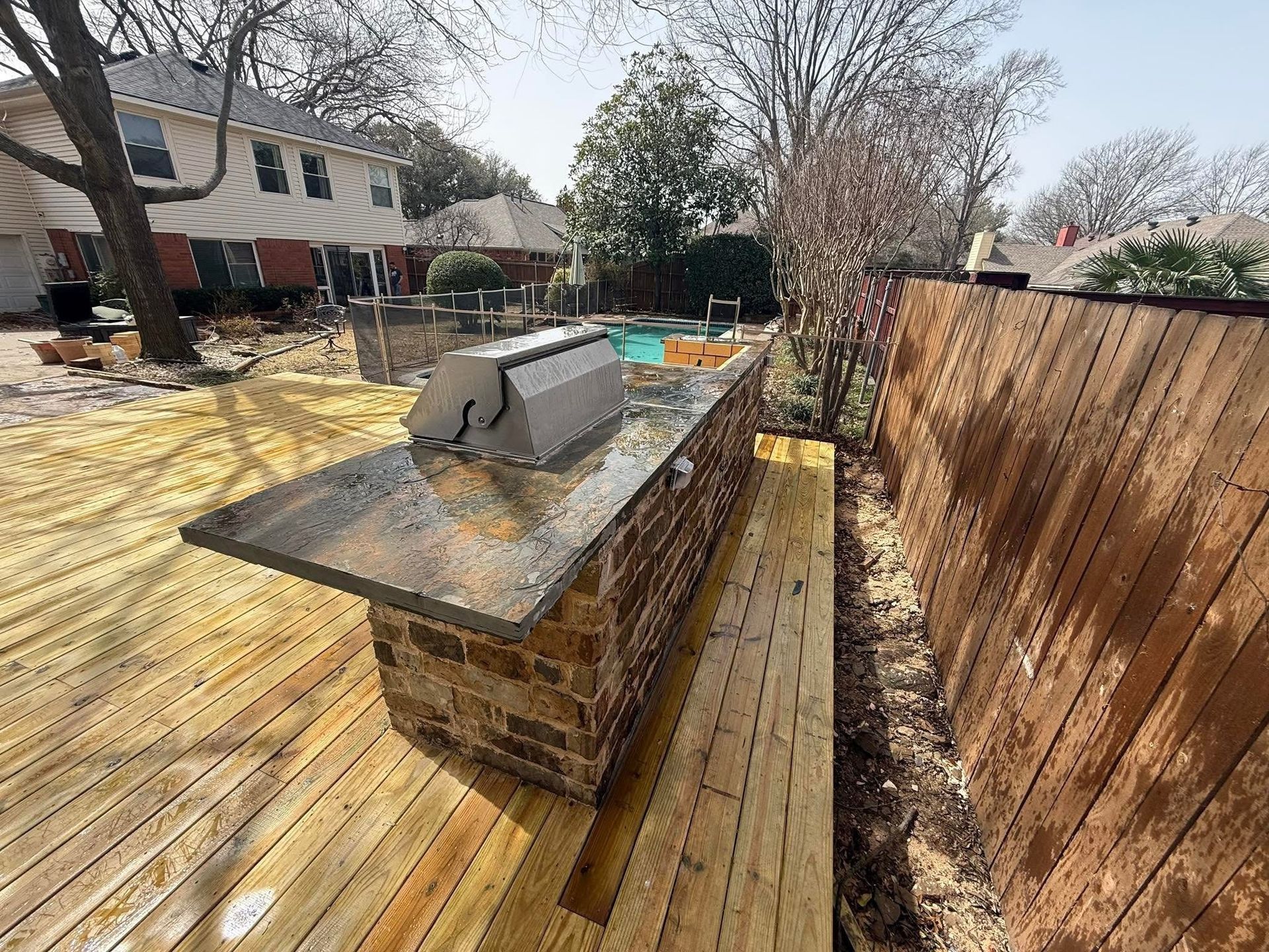 Backyard patio with wooden deck, brick outdoor grill island, and privacy fence on a sunny day