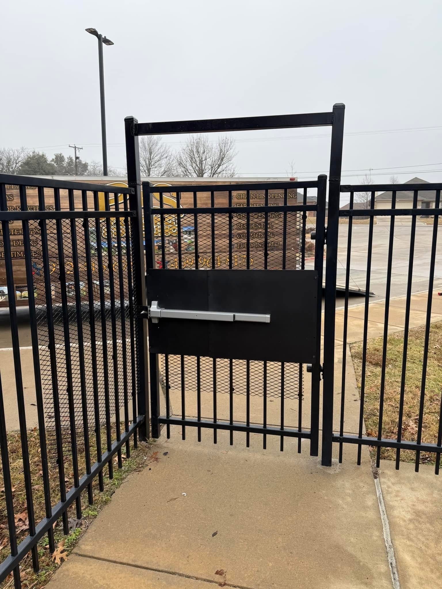 Black metal gate with a small dog door in a fenced outdoor area beside a paved path