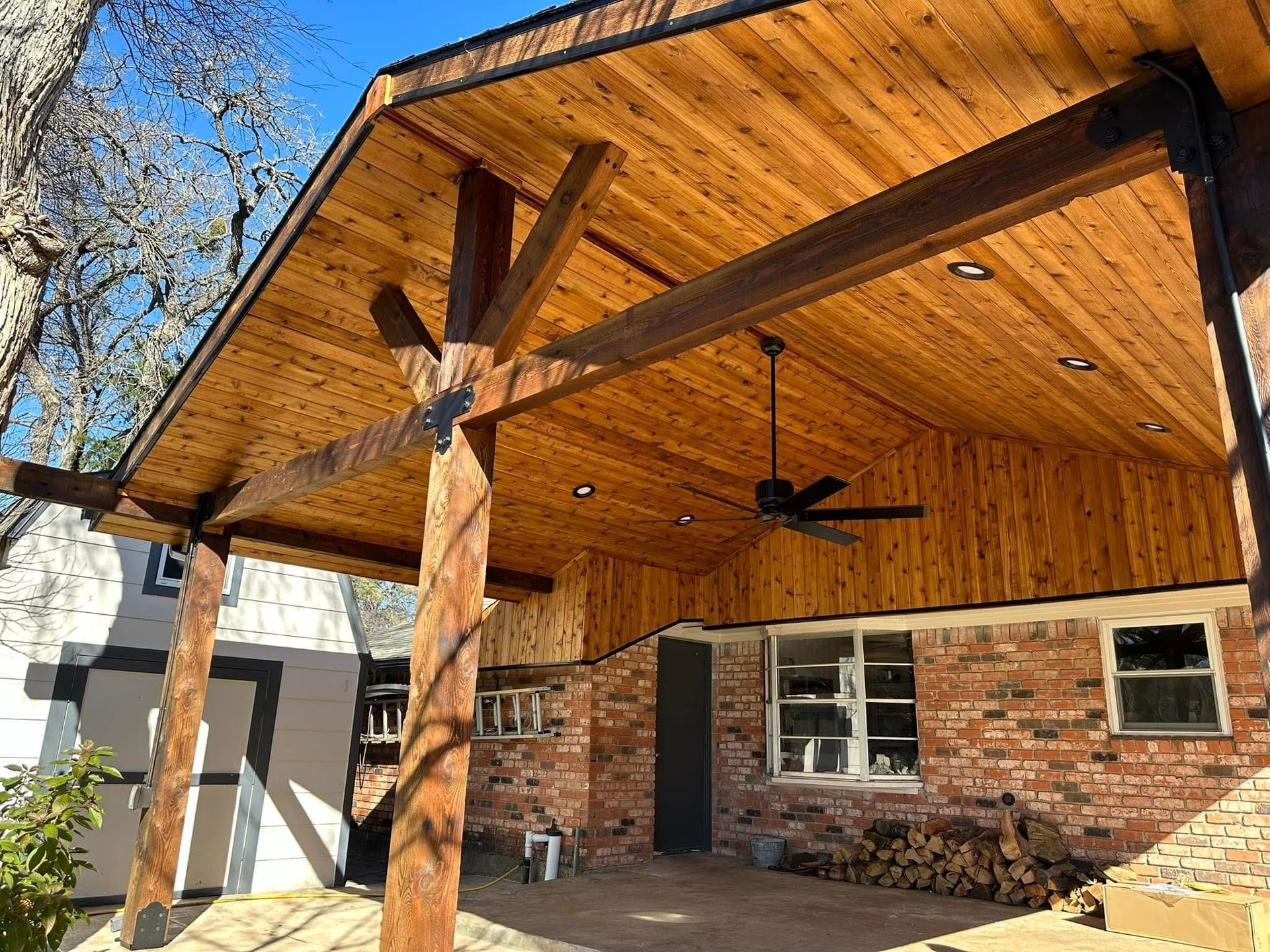 Wood-paneled patio with exposed beams, ceiling fans, brick wall, and stone fireplace under a covered roof