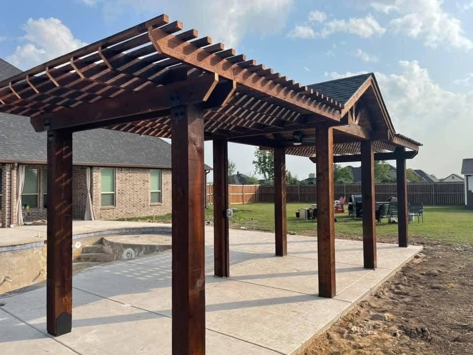 Wooden pergola over a concrete patio beside a brick house, with a yard and cloudy sky.