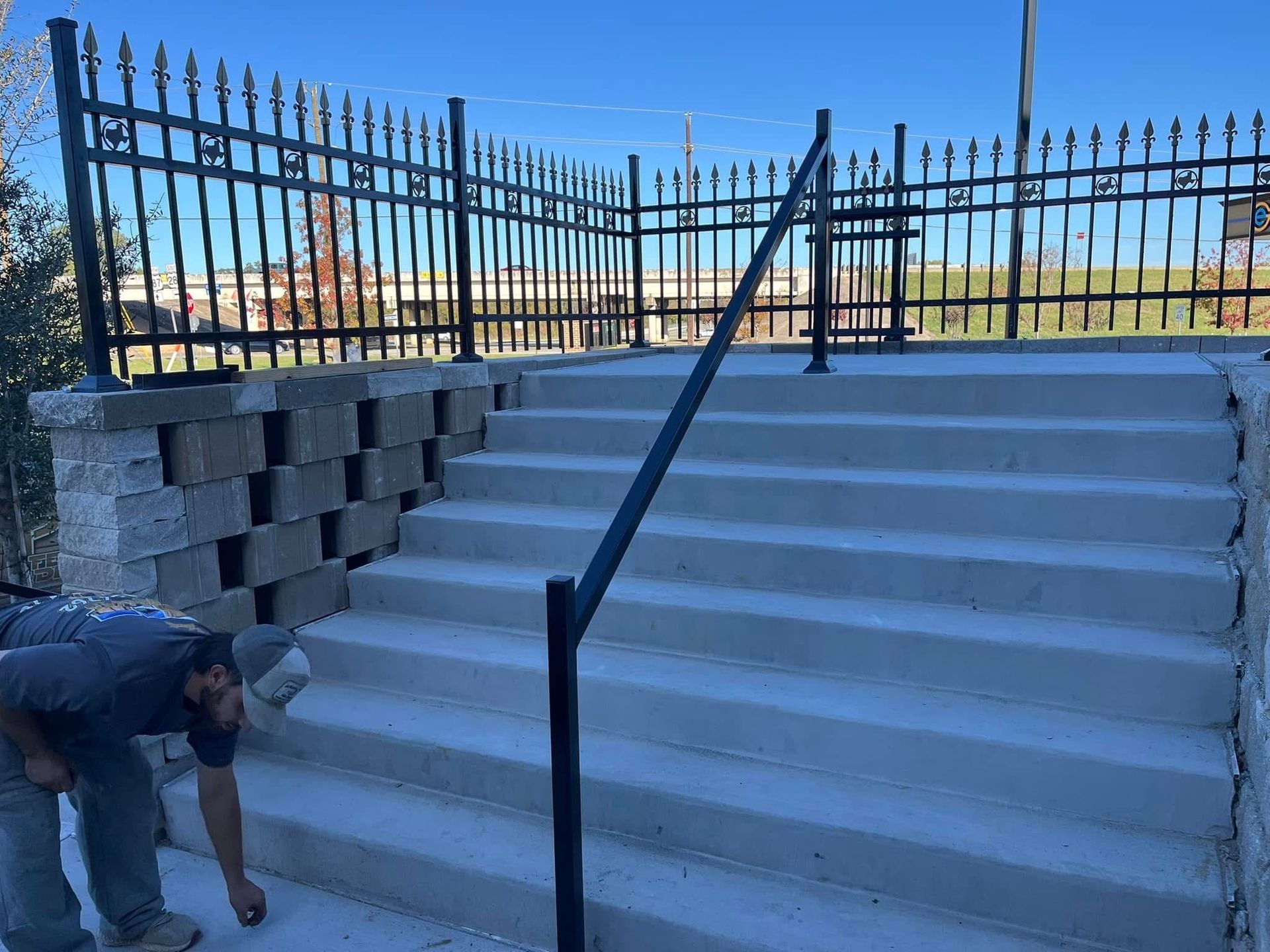 Concrete steps with black handrails beside a metal fence; a worker bends at the lower left.