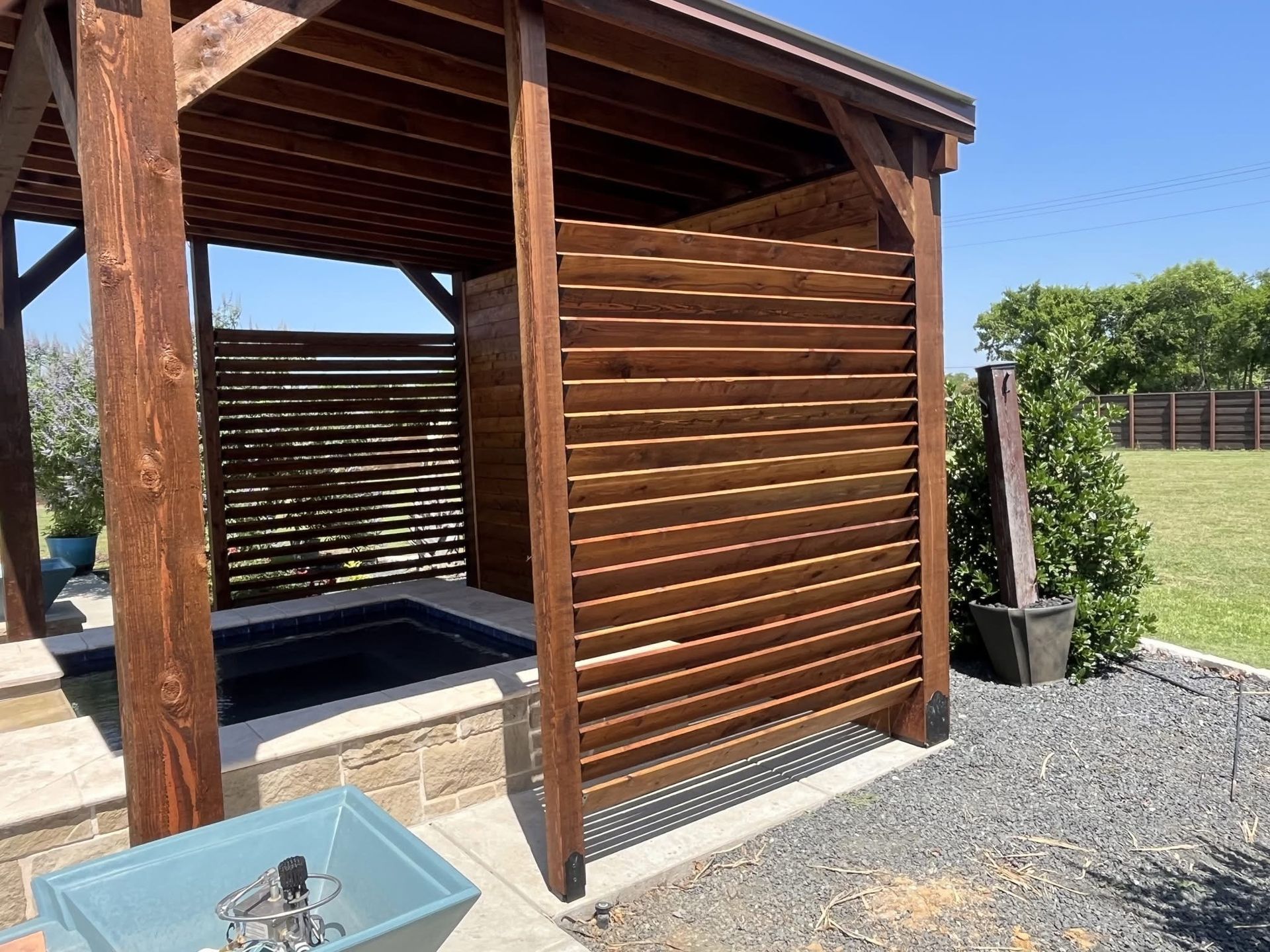 Wooden outdoor pergola with slatted privacy wall beside a gravel yard and grassy field under a clear sky