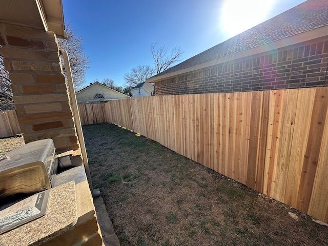 Narrow backyard walkway beside a wooden fence and brick house under a bright blue sky.