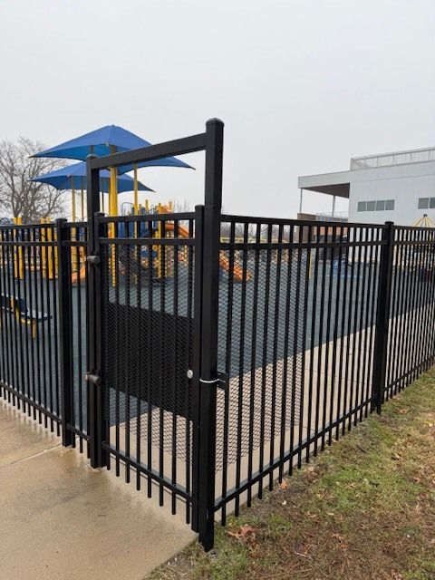 Black metal fence around a playground with blue shade structures beside a sidewalk and building