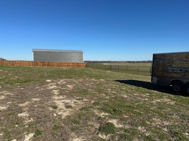 Grassy field with a large cylindrical metal tank under a clear blue sky, fence and trailer at right.