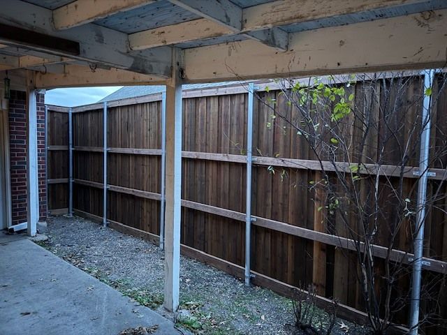 Covered backyard patio with wooden fence, white posts, and concrete floor next to a brick wall