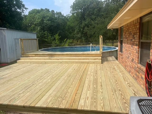Wooden deck with an above-ground pool beside a brick house and shed in a backyard.