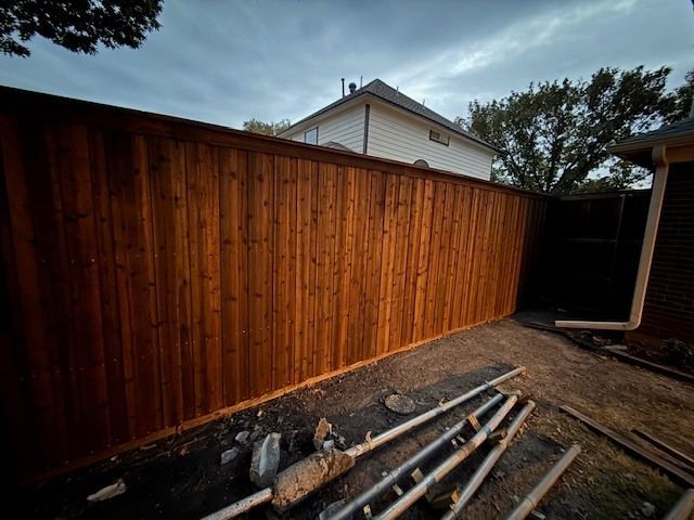 Wooden backyard fence beside a house, with metal pipes and debris piled along the ground.