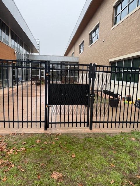 Black metal gate with a locked black panel between brick buildings and a patch of grass in front