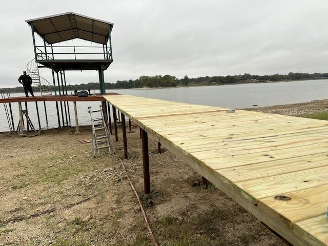 Wooden dock under construction extending toward a lake, with a ladder and small tower platform nearby.