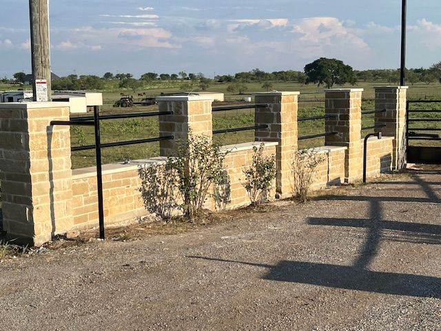 Stone-and-metal fence beside a gravel road with an open field and blue sky behind it